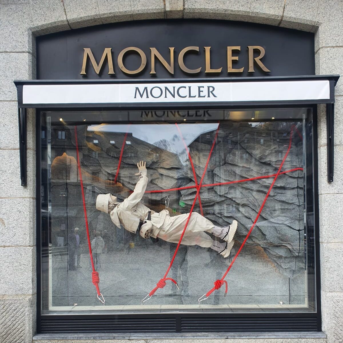Moncler window display in Milan with a mannequin suspended by red ropes over a mountain-themed backdrop.