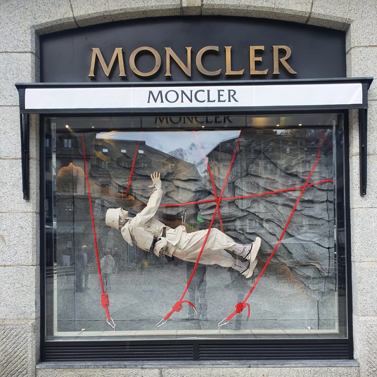 Home 4 Moncler window display in Milan with a mannequin suspended by red ropes over a mountain-themed backdrop.