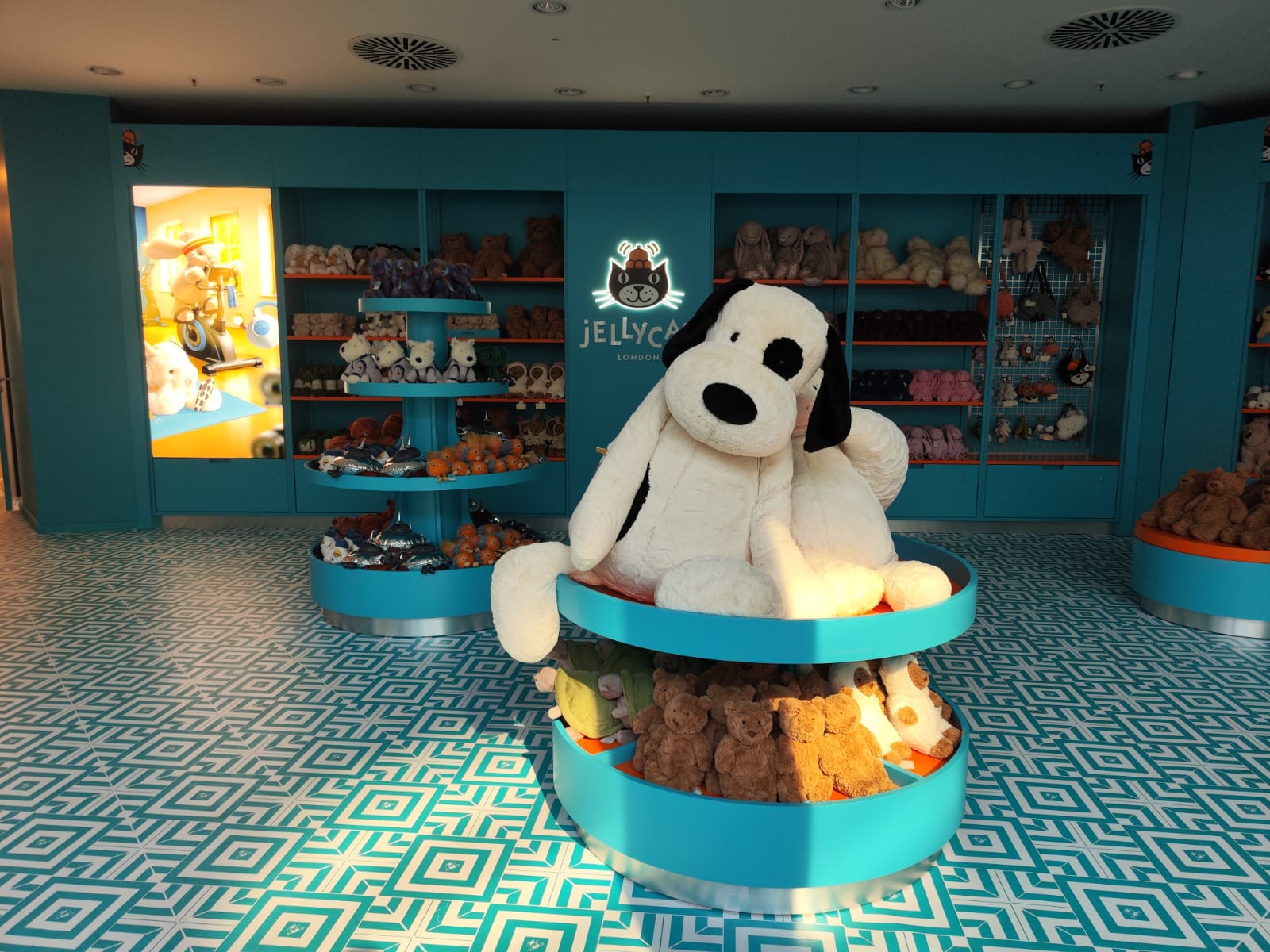 Panoramica dell'allestimento del negozio pop-up Jellycat a Stoccarda, con pareti azzurre, pavimentazione geometrica bianco-turchese ed espositori a isola ricchi di peluche.