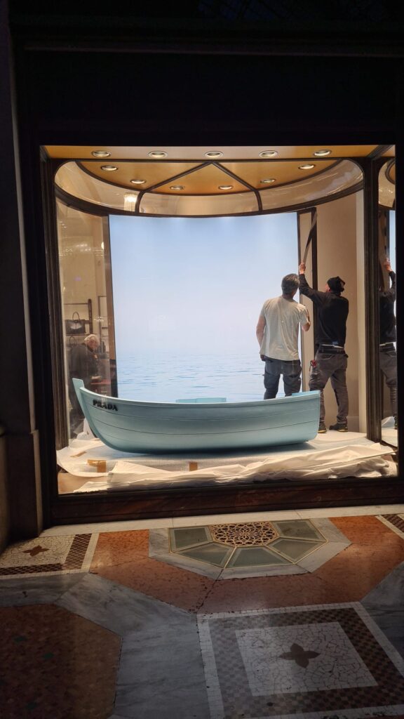 Home 9 Professional installers applying the backdrop and finishing the set design featuring a light blue boat in the Prada window display in Milan.