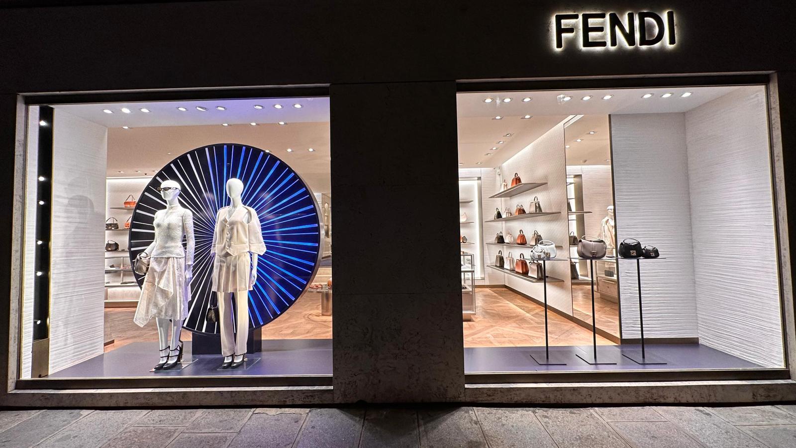 Facciata notturna di una boutique Fendi con due ampie vetrine illuminate: una a sinistra con manichini davanti a un grande pannello circolare a raggiera blu e una a destra con borse su piantane