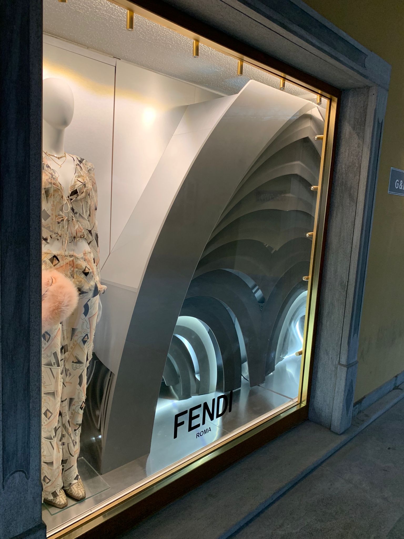 Scorcio angolare di una vetrina Fendi a Courmayeur, con un manichino in abito stampato di fianco a una maestosa struttura prospettica ad archi sovrapposti illuminati a LED