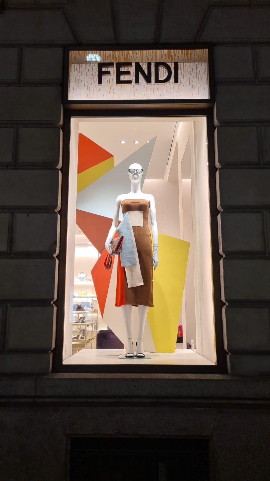 Vetrina Fendi a Milano con manichino che indossa un abito marrone e accessori, posizionato davanti a uno sfondo con pannelli geometrici colorati in arancione, giallo e grigio