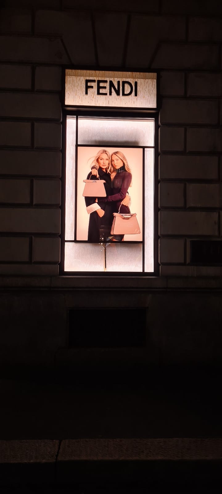 Insegna luminosa Fendi sopra una vetrina d'alta moda che mostra un poster pubblicitario di una campagna stagionale con due modelle che stringono delle borse rosa