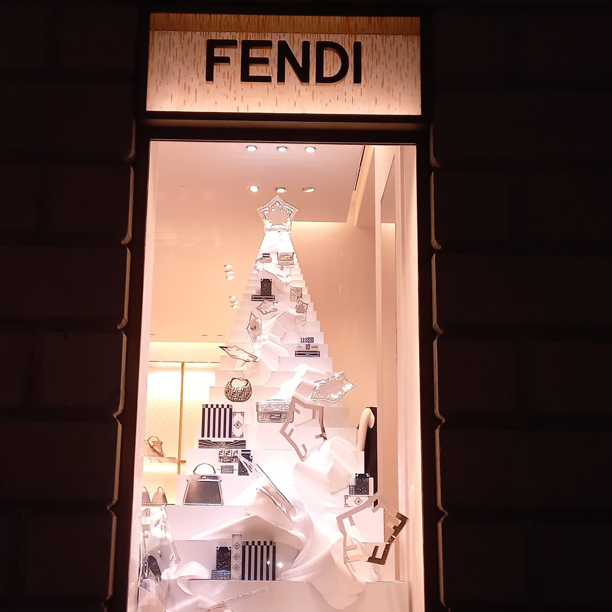 Allestimento di una vetrina natalizia Fendi a Milano, caratterizzato da una struttura scultorea bianca a forma di albero di Natale stilizzato con borse di lusso posizionate sui vari livelli.