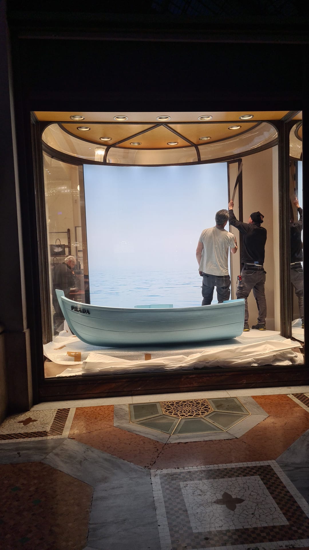 Professional installers applying the backdrop and finishing the set design featuring a light blue boat in the Prada window display in Milan.