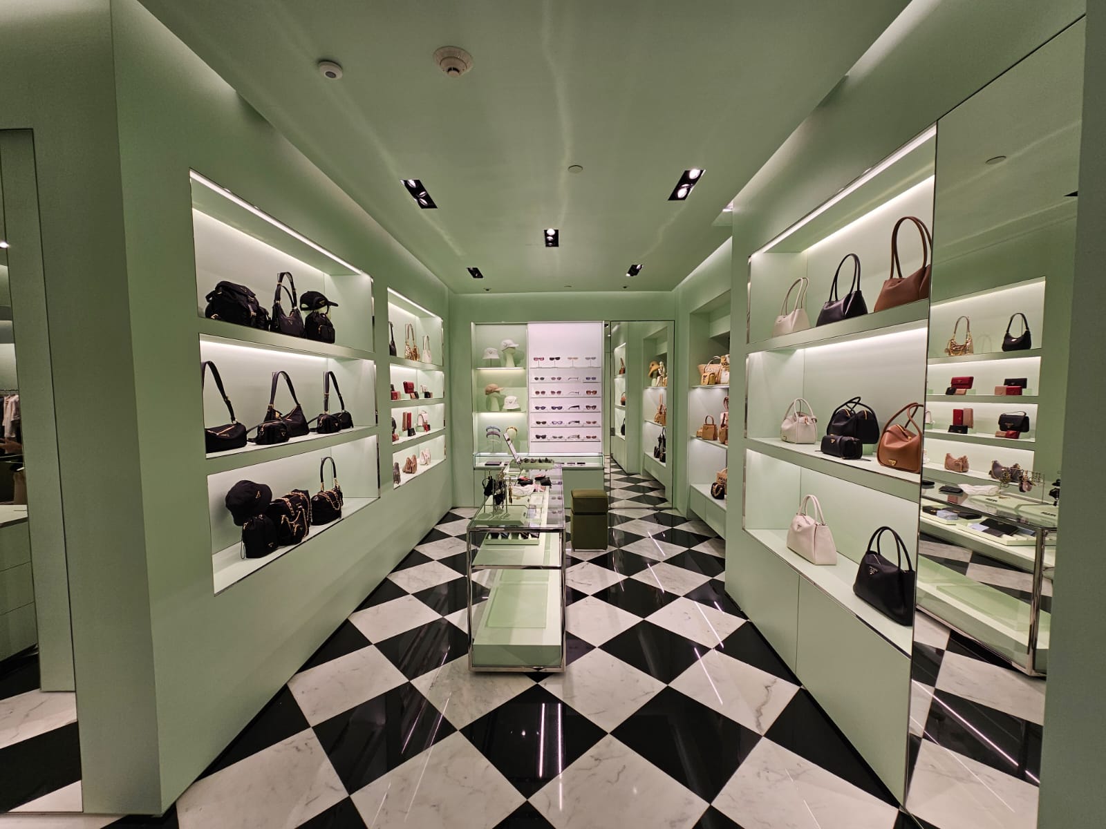 Allestimento interni del negozio Prada ad Aruba con pavimento a scacchi bianchi e neri, pareti verde pastello e scaffalature retroilluminate per borse.
