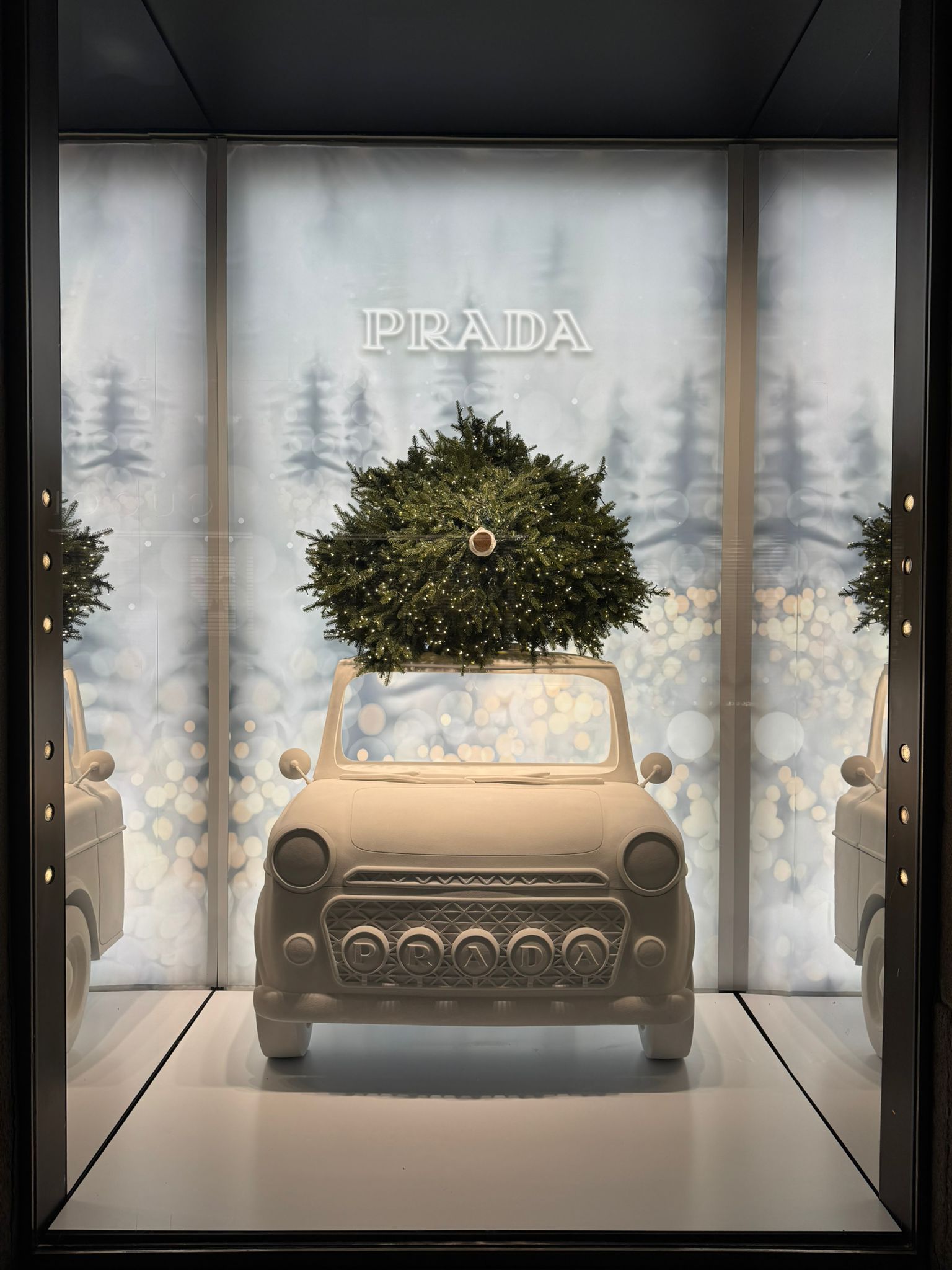 Vista frontale della vetrina di Natale Prada a Milano con fondale retroilluminato a tema foresta innevata e modello di auto d'epoca bianca custom.